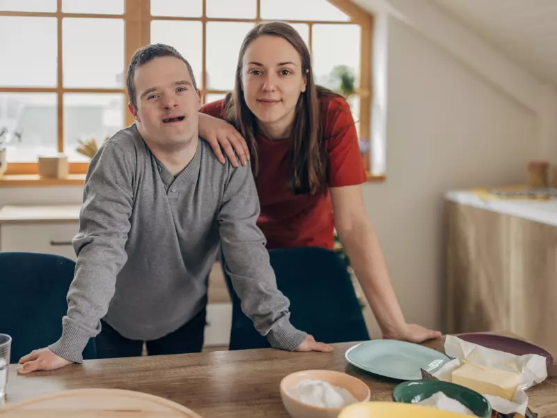 Disabled man with down syndrome smiling at camera with carer both leaning on table
