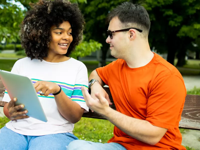 Disabled man with down syndrome sitting on park bench with support worker looking at iPad together